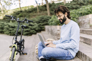 Smiling freelancer with bicycle using laptop sitting on steps - XLGF03007