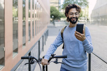 Smiling man with bicycle using phone in city - XLGF03016