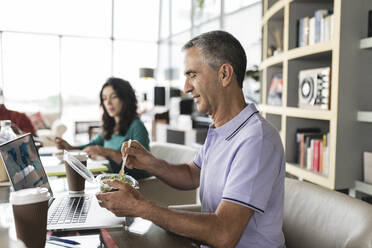 Happy businessman eating salad at desk in office - JCICF00188