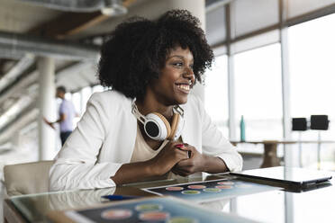 Afro-Geschäftsfrau mit drahtlosen Kopfhörern am Schreibtisch sitzend - JCICF00239