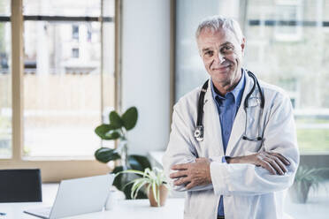 Happy doctor with arms crossed standing in office - UUF26645