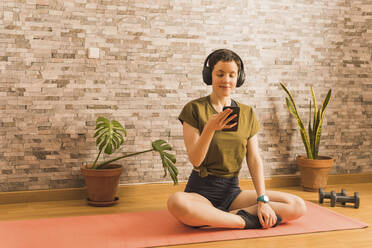 Woman using smart phone sitting on exercise mat - MGRF00680