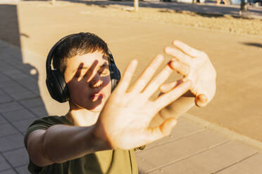 Frau mit drahtlosen Kopfhörern, die an einem sonnigen Tag die Augen mit den Händen abschirmt - MGRF00690