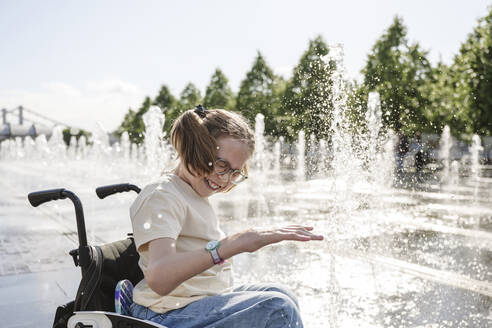 Fröhliches Mädchen mit Behinderung sitzt im Rollstuhl und spielt am Brunnen - EYAF01962