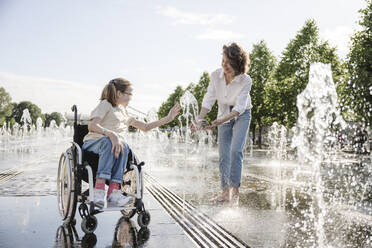 Mädchen im Rollstuhl, das mit seiner Mutter im Park am Wasser des Brunnens spielt - EYAF01968