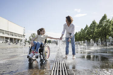 Mädchen im Rollstuhl spielt mit ihrer Mutter im Park am Brunnen - EYAF01969