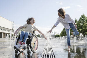 Frau und Tochter mit Behinderung spielen am Brunnen im Park an einem sonnigen Tag - EYAF01970