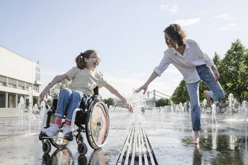 Woman and daughter with disability playing along fountain at park on sunny day - EYAF01970