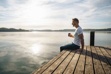 Mann sitzt mit einer Tasse Kaffee am Steg am See an einem sonnigen Tag - DIGF18126