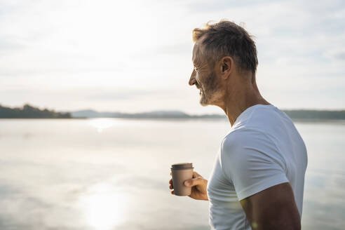 Mature man holding disposable coffee cup by lake on sunny day - DIGF18127
