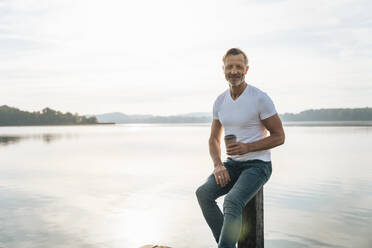 Happy mature man with disposable coffee cup sitting on post in front of lake - DIGF18131