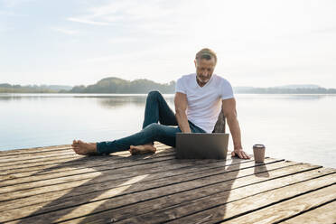 Freiberufler mit Einweg-Kaffeebecher am Laptop auf dem Bootssteg - DIGF18134