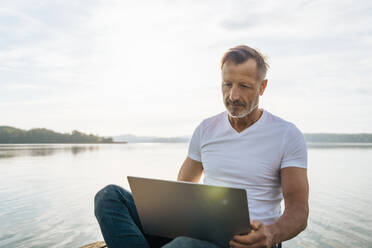 Älterer Freiberufler mit Laptop am Seeufer - DIGF18135