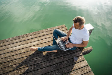 Freelancer working on laptop sitting at pier over lake - DIGF18138
