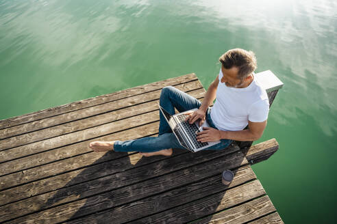 Freelancer working on laptop sitting at pier over lake - DIGF18138