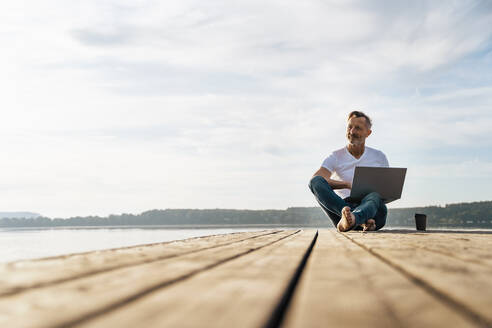 Nachdenklicher Freiberufler sitzt mit Laptop auf einem Pier über dem See - DIGF18139