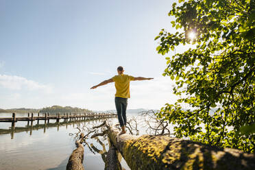 Man with arms outstretched balancing on fallen tree at lake - DIGF18151