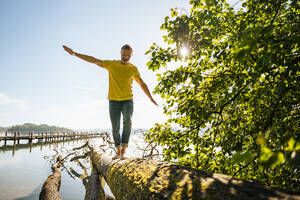 Mature man balancing on fallen tree at lakeshore - DIGF18153