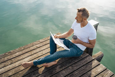 Man with newspaper and disposable coffee cup sitting on pier - DIGF18159