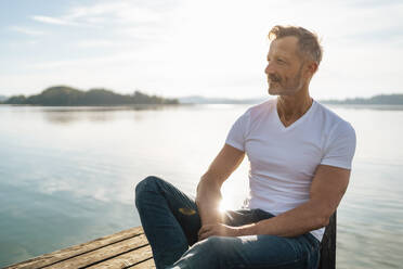 Smiling mature man sitting by lake on sunny day - DIGF18163