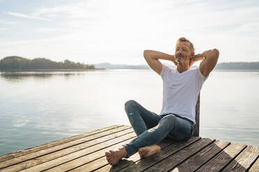 Älterer Mann entspannt sich mit den Händen hinter dem Kopf auf einem Pier am See - DIGF18165