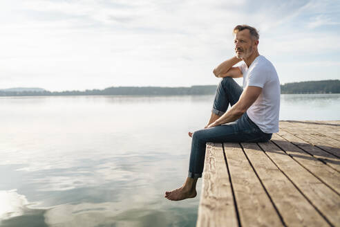 Thoughtful mature man sitting at the edge of pier by lake - DIGF18172