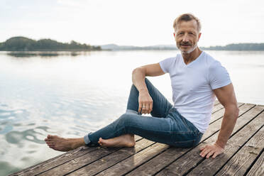 Smiling mature man sitting on jetty at lake - DIGF18174