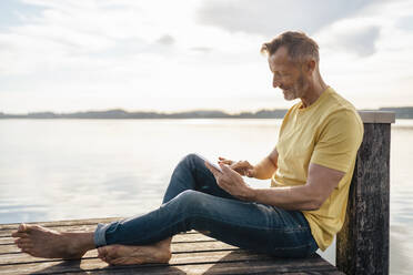 Älterer Mann mit Tablet-PC auf einem Pier am See sitzend - DIGF18182