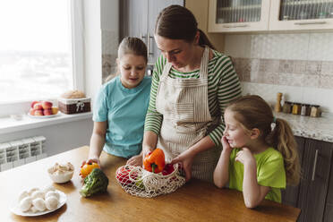 Girls looking at mother taking vegetables from mesh bag in kitchen - OSF00170
