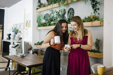 Smiling businesswoman sharing mobile phone with colleague in office - DCRF01257