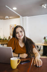 Lächelnde Geschäftsfrau sitzt mit Laptop und Kaffeetasse am Tisch im Büro - DCRF01279