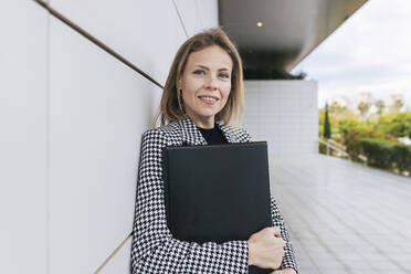 Smiling businesswoman holding file folder leaning on wall - JRVF02986
