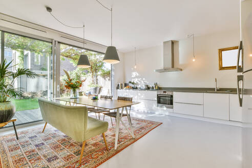 Lamps hanging over dining table with chairs and couch placed in contemporary spacious kitchen with white furniture and chrome appliances in modern house - ADSF35520