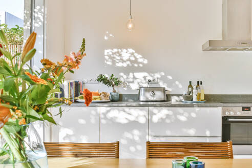 Lamps hanging over dining table with chairs in contemporary spacious kitchen with white furniture and chrome appliances in modern house - ADSF35521