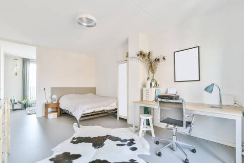 Bequemes Bett in der Nähe von Teppich in hellen geräumigen Schlafzimmer mit Sessel am Schreibtisch mit Lampe und dekorative Blumen zu Hause platziert - ADSF35524