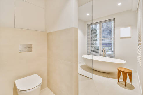 Keramische Toilette, die durch eine geflieste Trennwand von der Badewanne getrennt ist, in einem hellen Badezimmer eines modernen Hauses - ADSF35540