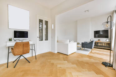 Desk with computer and chair located near white door and spacious living room in daytime at home - ADSF35552