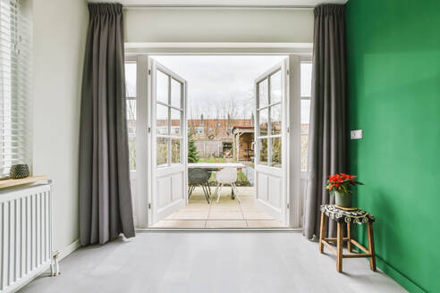 Potted plant placed on stool at green wall in spacious apartment near opened doors leading to terrace with table and chairs against cloudy sky - ADSF35604