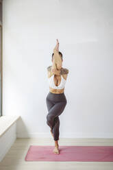 Woman practicing Garudasana on exercise mat in front of wall at yoga studio - MTBF01216