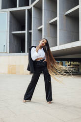 Confident young businesswoman with long braided brown hair standing in front of office building - JRVF03019
