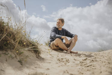 Glücklicher reifer Mann sitzt auf Sand am Strand - MFF09174