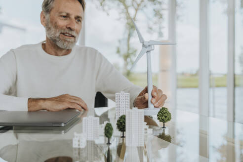 Smiling mature man with wind turbine model sitting at table - MFF09199