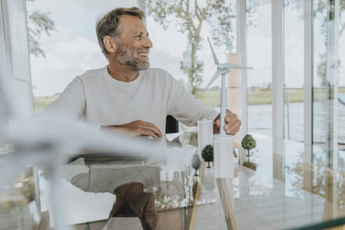 Happy mature man with wind turbine model sitting at home - MFF09200