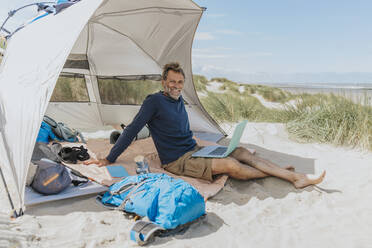 Glücklicher Freiberufler mit Laptop im Zelt am Strand an einem sonnigen Tag - MFF09220