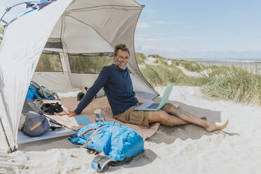 Happy freelancer with laptop sitting in tent at beach on sunny day - MFF09220