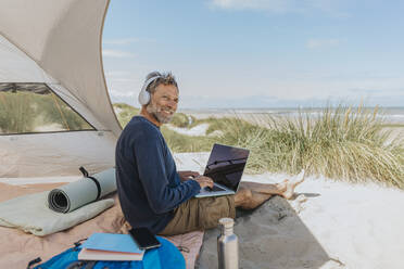 Happy freelancer wearing wireless headphones sitting at beach - MFF09221