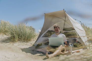 Glücklicher Freiberufler mit Laptop im Zelt sitzend an einem sonnigen Tag - MFF09230