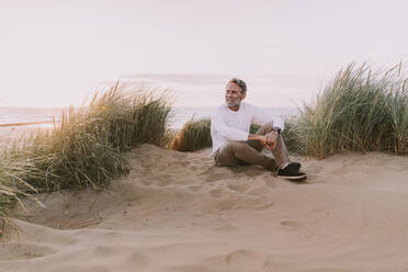 Glücklicher Mann sitzt auf einer Sanddüne am Strand - MFF09238