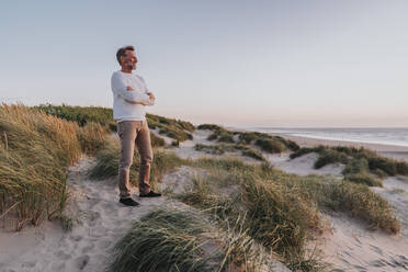 Happy man with arms crossed standing on sand dune at beach - MFF09239