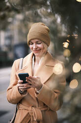Happy woman wearing knit hat using mobile phone - AGOF00277